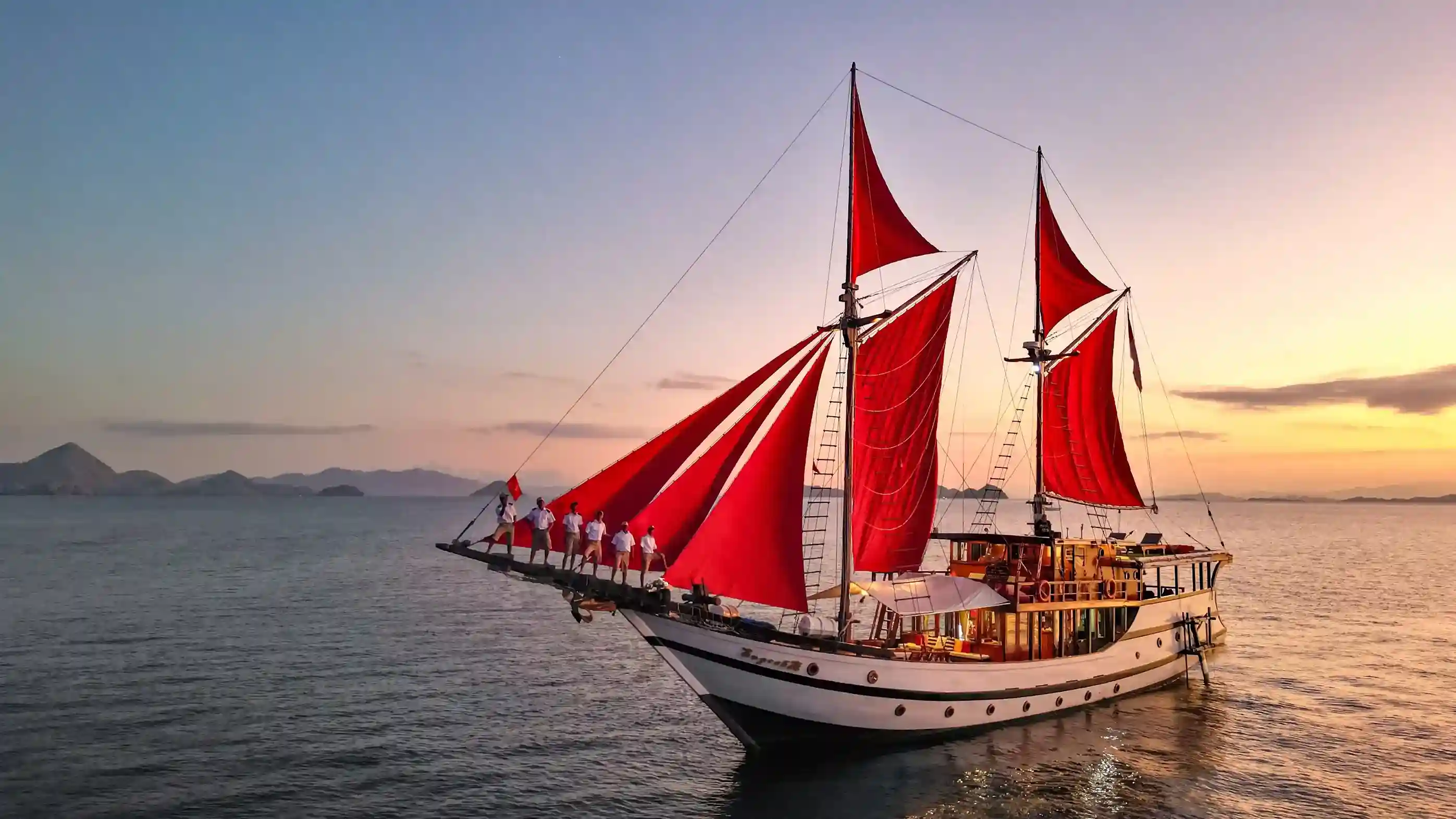 A phinisi with red sails underway at sunset